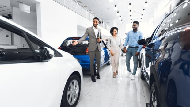 Car Selling Business. Manager Showing Luxury Automobile To Afro Spouses In Automobile Dealership Center. Panorama