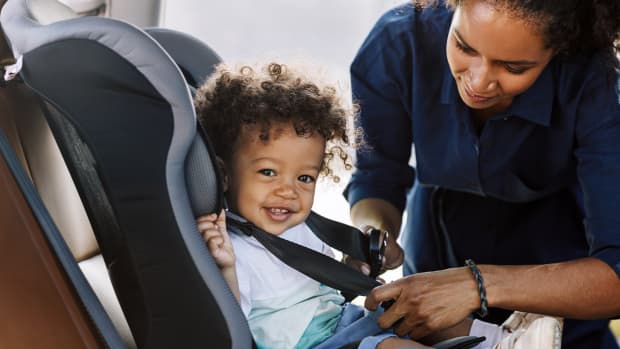 Side view of a happy little boy looking at camera while his mother buckling him in a car seat